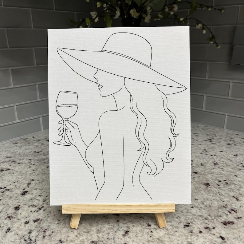 May include: A white canvas featuring a black line art illustration of a person with long wavy hair wearing a wide-brimmed sun hat and holding a stemmed wine glass. The artwork is displayed on a small wooden easel stand, showcasing a simple profile.