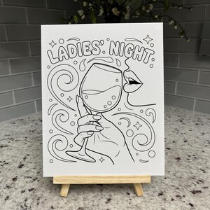 May include: A white canvas featuring a black line art illustration of a hand holding a wine glass, with the text LADIES' NIGHT above. The design includes swirling decorative lines, small stars, and a partial profile of a face with lips.