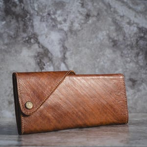May include: A brown leather wallet with a button closure. The wallet has a textured surface and a diagonal flap. The stitching is visible along the edges. The wallet is rectangular and appears to be new.