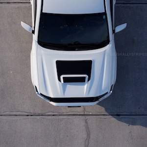 May include: A white pickup truck with a black hood scoop. The truck is parked on a concrete surface.