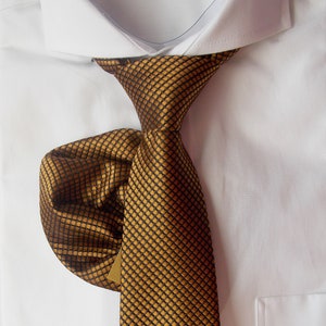 Może przedstawiać: Zbliżenie złotej i czarnej wzorzystej krawata zawiązanego i włożonego w białą koszulę. Krawat ma powtarzający się kwadratowy wzór. Koszula ma kieszeń po prawej stronie.