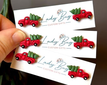 Puede incluir: Pendientes de camión rojo con un árbol de Navidad verde en la caja. Los pendientes están en tarjetas blancas con el texto "Lady Bug Custom Gifts". Un accesorio festivo para las fiestas.
