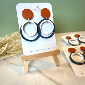 May include: Handmade polymer clay earrings. Each earring features a rust-colored disc, a white circle, and a navy blue circle. The earrings are displayed on a white card with the text "Lady Bug Custom Gifts" and "Handmade Polymer Clay Earrings".