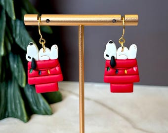 Puede incluir: Un par de pendientes con temática de Snoopy. Cada pendiente presenta una caseta roja para perros con Snoopy acostado encima. La caseta tiene una cadena de luces de colores. Los pendientes cuelgan de ganchos dorados.