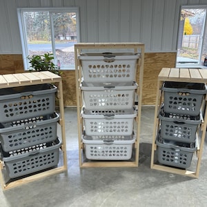 May include: Three wooden laundry basket organizers. The organizers have a natural wood finish and feature multiple gray or white plastic laundry baskets. The baskets are stacked within the wooden frames, offering a practical storage solution for laundry rooms.