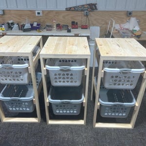 May include: Three wooden storage units with light-coloured tops and frames. Each unit has two grey plastic laundry baskets. The units are designed for laundry organisation and storage.