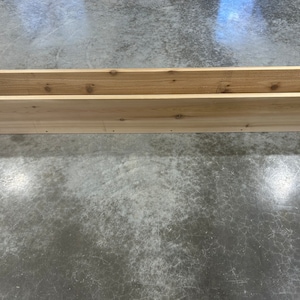 Op de afbeelding: Een lange, rechthoekige houten plantenbak gemaakt van natuurlijk cederhout. De plantenbak is leeg en staat op een betonnen vloer.