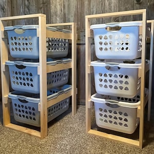 May include: Two wooden laundry sorters with light blue plastic baskets. Each sorter has three baskets stacked within a light-colored wooden frame. The baskets have a perforated design for ventilation, and the frames are made of unfinished wood.