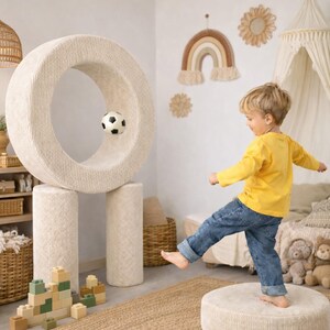Op de afbeelding: Een kind in een geel shirt en een blauwe spijkerbroek schopt een voetbal door een grote, beige, cirkelvormige speelstructuur. De structuur wordt ondersteund door twee beige cilinders. Een kleine, zwart-witte voetbal hangt in de lucht. Een beige, ronde poef staat op de grond.