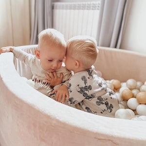 Könnte beinhalten: Zwei Kleinkinder sitzen in einem weißen Ballbecken. Das Kleinkind links trägt ein weißes Shirt mit einem braunen und weißen Muster. Das Kleinkind rechts trägt ein weißes Shirt mit einem schwarzen und weißen Mickey-Mouse-Muster. Das Ballbecken ist mit weißen und beigefarbenen Bällen gefüllt.