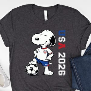 May include: Dark gray t-shirt featuring Snoopy in a soccer uniform with a soccer ball. The shirt has the text "USA 2026" in red, white, and blue. The image also includes blue jeans and white sneakers.