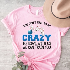 May include: Pink t-shirt with a bowling ball and pins graphic. The text on the shirt reads "You don't have to be crazy to bowl with us, we can train you."