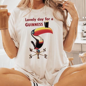 May include: Cream-colored t-shirt featuring a toucan design with the text "Lovely day for a Guinness." The toucan has a colorful beak and is perched on a weather vane. The person is holding a glass jar with a straw.