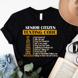 May include: Black t-shirt with a white graphic that says "Senior Citizen Texting Code" and lists common texting abbreviations and their meanings. The abbreviations are in yellow text.