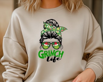May include: A beige crewneck sweatshirt featuring a graphic print of a messy bun hairstyle with a green patterned headband, aviator sunglasses reflecting a Grinch character in a Santa hat, and the text "GRINCH life" in green and black lettering.