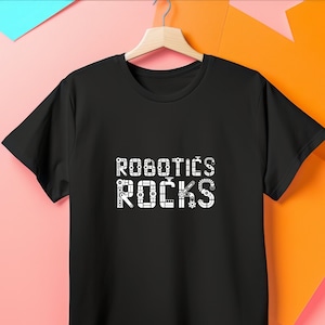 May include: Black t-shirt with the words "ROBOTICS ROCKS" in white, stylized with mechanical and robotic elements. The shirt is hanging on a wooden hanger against a colorful background.