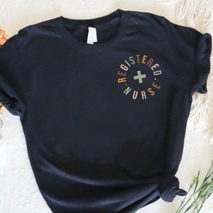 May include: A black short-sleeve t-shirt featuring a circular graphic on the left chest. The text reads REGISTERED NURSE in a circular arrangement with a light green cross symbol in the center. The lettering uses alternating muted earth tones.