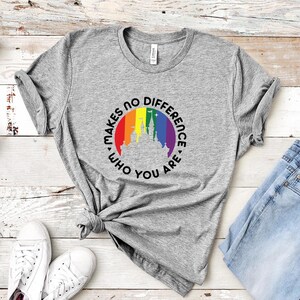 May include: A heather gray short-sleeve t-shirt featuring a circular graphic with a rainbow-colored background and a white silhouette of a castle. The text "MAKES NO DIFFERENCE WHO YOU ARE" is printed in black capital letters around the circle.