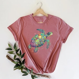May include: A pink t-shirt with a colorful mosaic sea turtle design. The turtle is facing right and has a green, blue, and yellow shell.