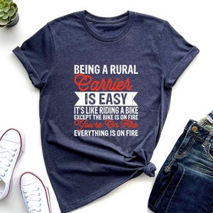 May include: A dark heather navy blue short-sleeve t-shirt with white and red text that reads: BEING A RURAL Carrier IS EASY IT'S LIKE RIDING A BIKE EXCEPT THE BIKE IS ON FIRE You're On Fire EVERYTHING IS ON FIRE. The shirt has a crew neckline.