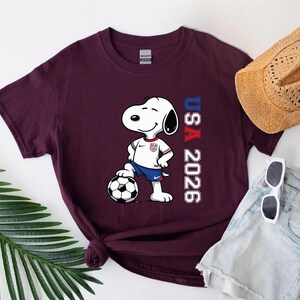 May include: Burgundy t-shirt featuring Snoopy in a soccer uniform, standing on a soccer ball. The shirt has the text "USA 2026" in red and blue. A straw hat, sunglasses, and denim shorts are also visible.