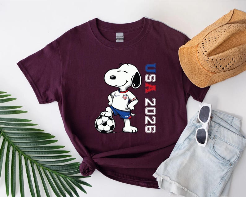 May include: Burgundy t-shirt featuring Snoopy in a soccer uniform, standing on a soccer ball. The shirt has the text "USA 2026" in red and blue. A straw hat, sunglasses, and denim shorts are also visible.