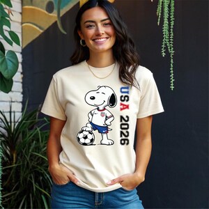 May include: Cream-colored t-shirt featuring a cartoon Snoopy character in a soccer uniform, standing on a soccer ball. The shirt has the text "USA 2026" printed vertically in red, white, and blue.