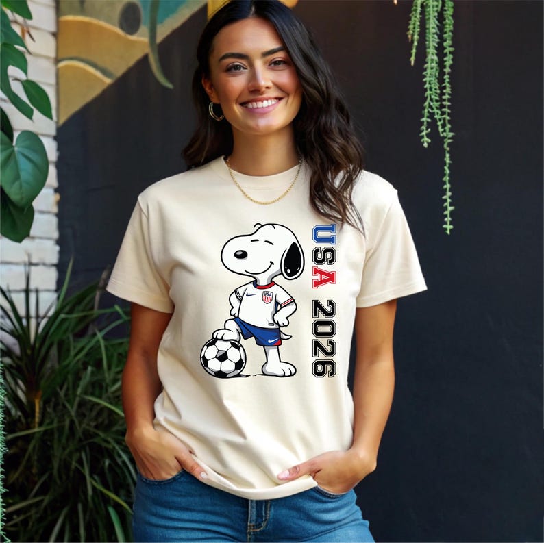 May include: Cream-colored t-shirt featuring a cartoon Snoopy character in a soccer uniform, standing on a soccer ball. The shirt has the text "USA 2026" printed vertically in red, white, and blue.