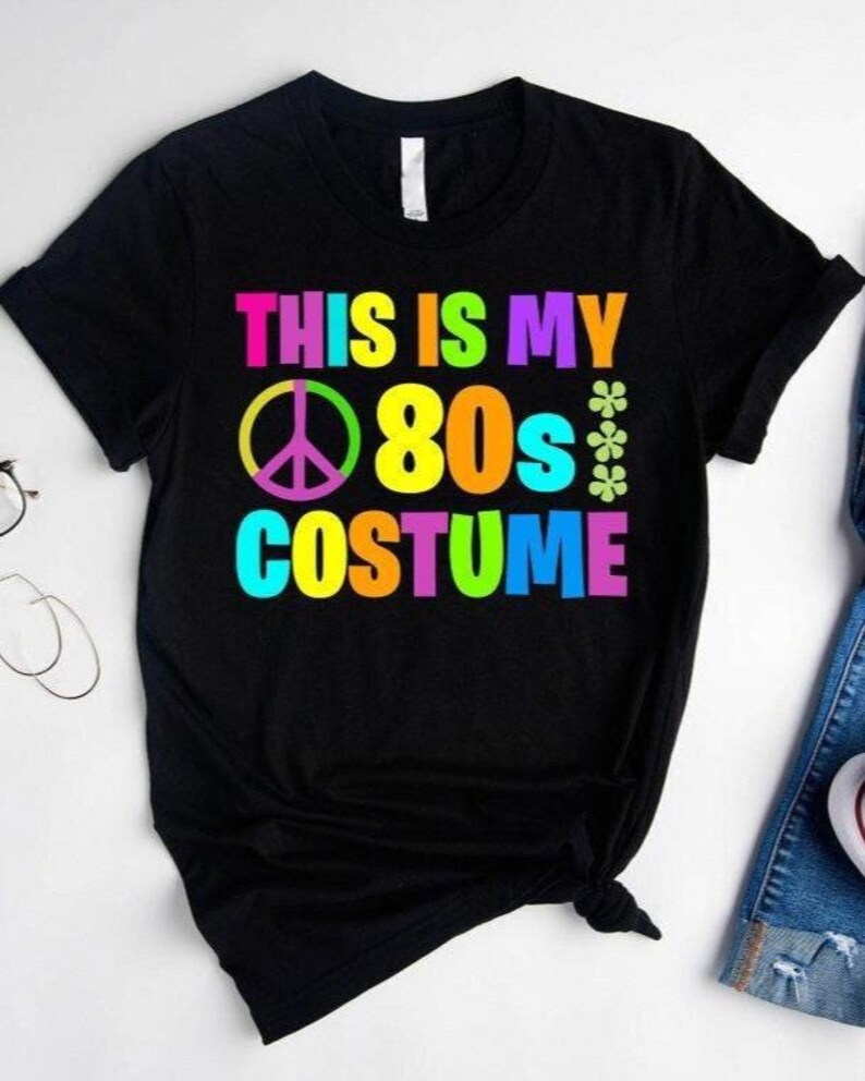 May include: Black t-shirt with the text "This is my 80s Costume" in rainbow colors. A peace sign is included in the design.