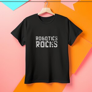 May include: A black short-sleeve t-shirt featuring the white text ROBOTICS ROCKS printed on the chest. The letters are stylized with mechanical components, including gears, joints, and pipes, creating a technical and industrial aesthetic.