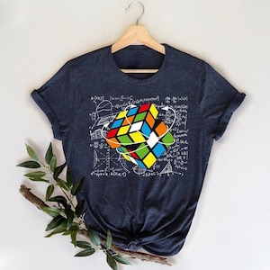May include: A dark charcoal gray short-sleeve t-shirt featuring a graphic print of a colorful Rubik's cube surrounded by white mathematical equations and geometric diagrams. The shirt has a crew neckline and is displayed on a wooden hanger.
