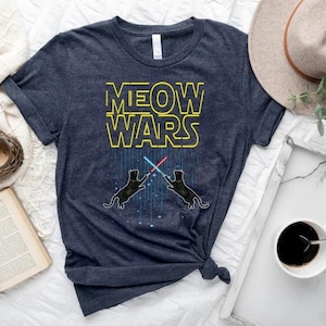 May include: A dark heather navy blue short-sleeve t-shirt featuring the yellow text MEOW WARS above a graphic of two black cats standing on their hind legs, each holding a glowing red or blue light saber, with vertical blue light streaks.