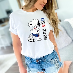 May include: White t-shirt featuring a cartoon Snoopy character in a soccer uniform, standing next to a soccer ball. The shirt has the text "USA 2026" printed vertically in red, white, and blue.