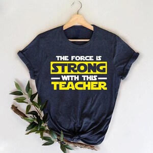 May include: A dark heather navy blue short-sleeve t-shirt featuring the white and yellow text "THE FORCE IS STRONG WITH THIS TEACHER" printed on the front. The shirt is displayed on a wooden hanger and has a crew neckline and a standard fit.