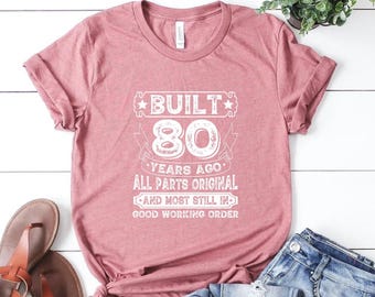 May include: A dusty rose short-sleeved t-shirt featuring white text that reads BUILT 80 YEARS AGO ALL PARTS ORIGINAL AND MOST STILL IN GOOD WORKING ORDER. The shirt is displayed alongside a pair of blue denim jeans and brown sandals.