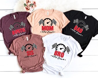 May include: Five short-sleeve t-shirts in maroon, peach, black, mauve, and white. Each shirt features a graphic of a speedometer flanked by two checkered flags, with red text reading AUNTIE, MOM, DAD, SIS, or BRO above the words Pit Crew.