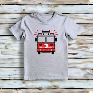 May include: A light gray short-sleeve t-shirt featuring a graphic of a red and black fire truck on the front. The truck design includes the number 3 in white, the text FIRE DEPT in white, and red light rays emanating from the top of the vehicle.