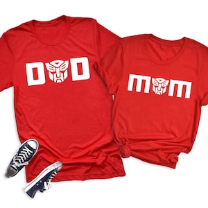 May include: Two bright red short-sleeved t-shirts. The left shirt features the white text DAD with a Transformer logo replacing the letter A. The right shirt features the white text MOM with a Transformer logo replacing the letter O.