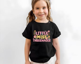 May include: A black short-sleeved t-shirt featuring a graphic print with the text "LITTLE MISS THREENAGER" in pink and yellow lettering, surrounded by colorful illustrations of various small flowers and foliage in shades of pink, yellow, and orange.
