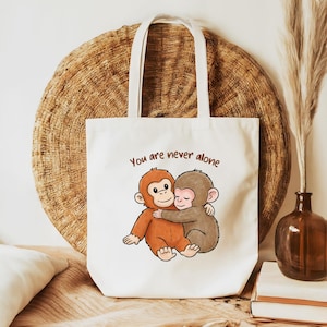 May include: A white canvas tote bag featuring a cartoon illustration of an orange orangutan and a grey monkey hugging. Above the illustration, the text "You are never alone" is printed in brown. The bag has two white fabric shoulder straps.