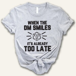 May include: A light gray short-sleeve t-shirt featuring black text that reads "WHEN THE DM SMILES IT'S ALREADY TOO LATE" with a graphic of a twenty-sided die showing the number one in the center, all printed in a distressed, textured font style.