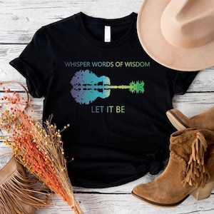 May include: A black short-sleeve t-shirt featuring a graphic of a guitar silhouette filled with a gradient of purple, blue, and green colors. The text "WHISPER WORDS OF WISDOM" appears above the guitar and "LET IT BE" is printed below it.