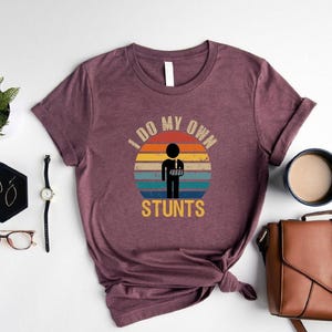 May include: A maroon t-shirt with a vintage-style graphic of a person with a broken arm and the text "I do my own stunts".