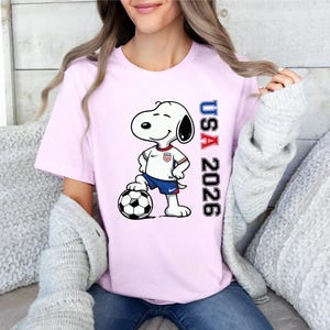 May include: A light pink t-shirt featuring Snoopy in a soccer uniform, standing next to a soccer ball. The shirt has the text "USA 2026" in red and blue. The design is centered on the front of the shirt.