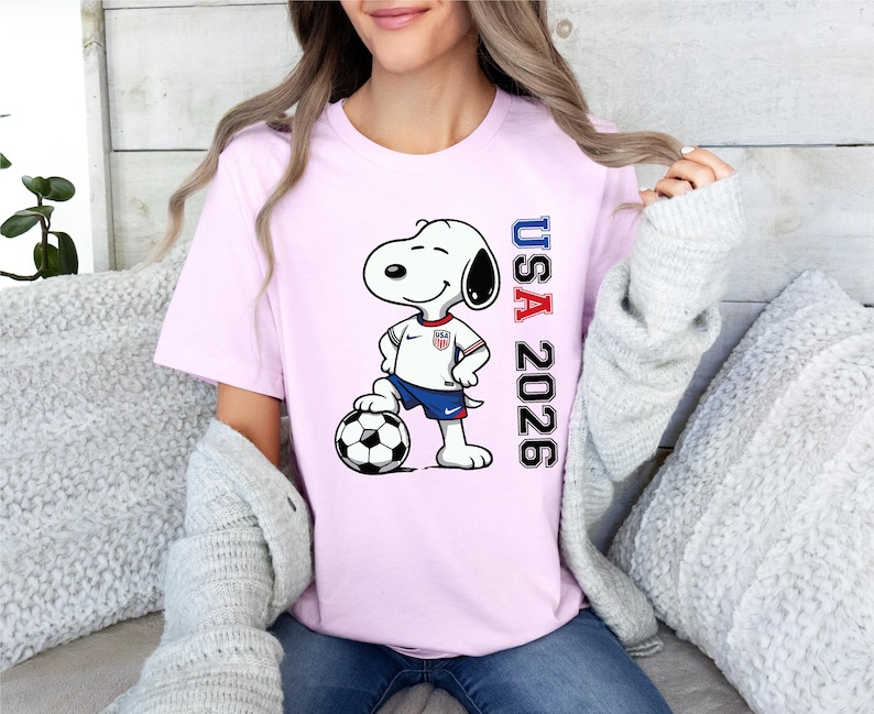 May include: A light pink t-shirt featuring Snoopy in a soccer uniform, standing next to a soccer ball. The shirt has the text "USA 2026" in red and blue. The design is centered on the front of the shirt.