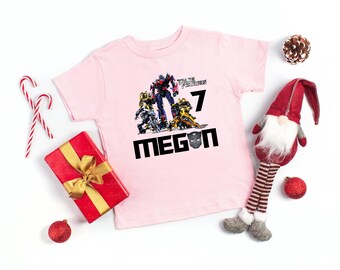 May include: A light pink short-sleeved t-shirt featuring a graphic of Transformers characters, the number 7, and the name MEGAN with a black robot mask icon replacing the letter A. The design is printed in the center of the shirt.