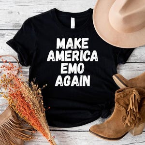 May include: Black t-shirt with white text that reads "Make America Emo Again". The shirt is laid flat on a wooden surface with a brown hat and brown boots.