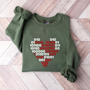 May include: An olive green crewneck sweatshirt featuring a heart-shaped graphic composed of binary code. The binary digits are printed in alternating white and red colors, forming a distinct heart pattern centered on the front of the garment.