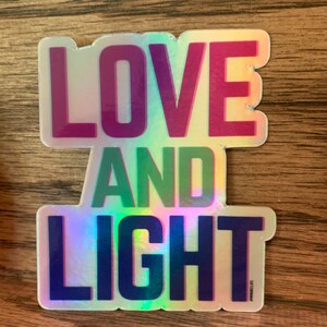 May include: A holographic sticker with the words "LOVE AND LIGHT" in rainbow colors. The sticker has a slightly iridescent finish.