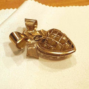 May include: A gold-toned metal brooch featuring a bow attached to a heart-shaped cage pendant. The heart cage is constructed with a grid-like frame and contains several small, round, off-white pearls visible through the open metal lattice design.